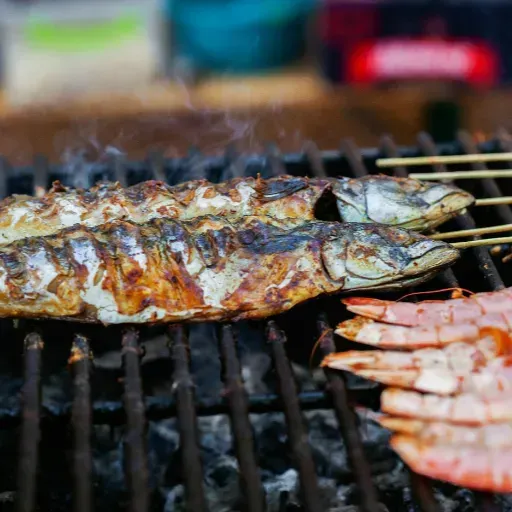 Gegrilde vis en garnalen aan spiesjes boven hete houtskool, met zichtbare grillstrepen.