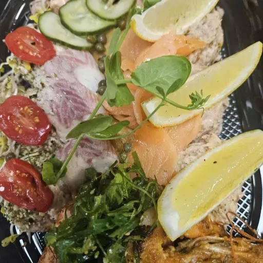 Sushischotel met zalm, komkommer, tomaten, zeewiersalade en citroenpartjes op een donker bord.
