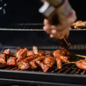 Een hand smeert barbecuesaus over sissend gegrild vlees op een zwarte grill.
