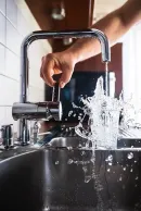A hand adjusts a chrome kitchen faucet as water splashes forcefully into a glass inside a stainless steel sink.