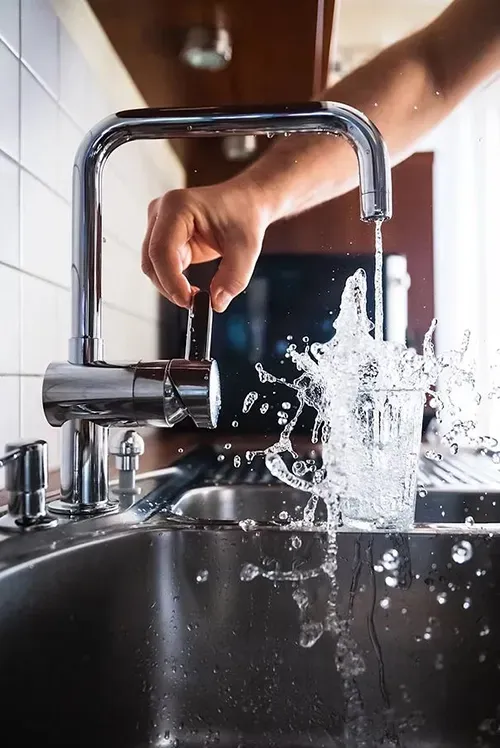 A hand adjusts a chrome kitchen faucet as water splashes forcefully into a glass inside a stainless steel sink.
