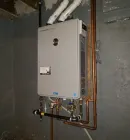 A grey, wall-mounted tankless water heater installed in a basement, connected to white exhaust pipes and copper plumbing.