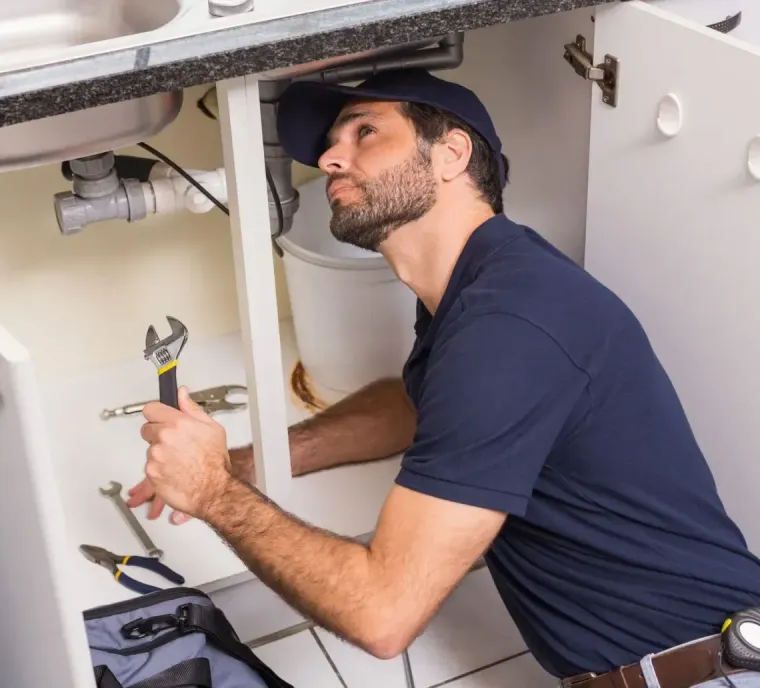 A person in a blue shirt and cap works under a kitchen sink, holding an adjustable wrench while checking the pipes.