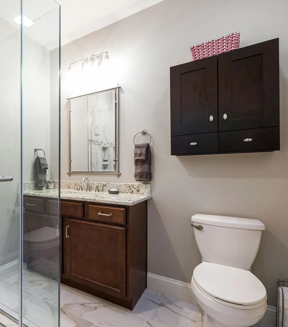 A bright bathroom featuring a walk-in glass shower, dark wood vanity, toilet, and mounted dark cabinet on a gray wall.
