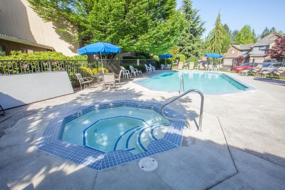 Outdoor swimming pool with a hot tub, lounge chairs, and blue umbrellas in a sunny courtyard