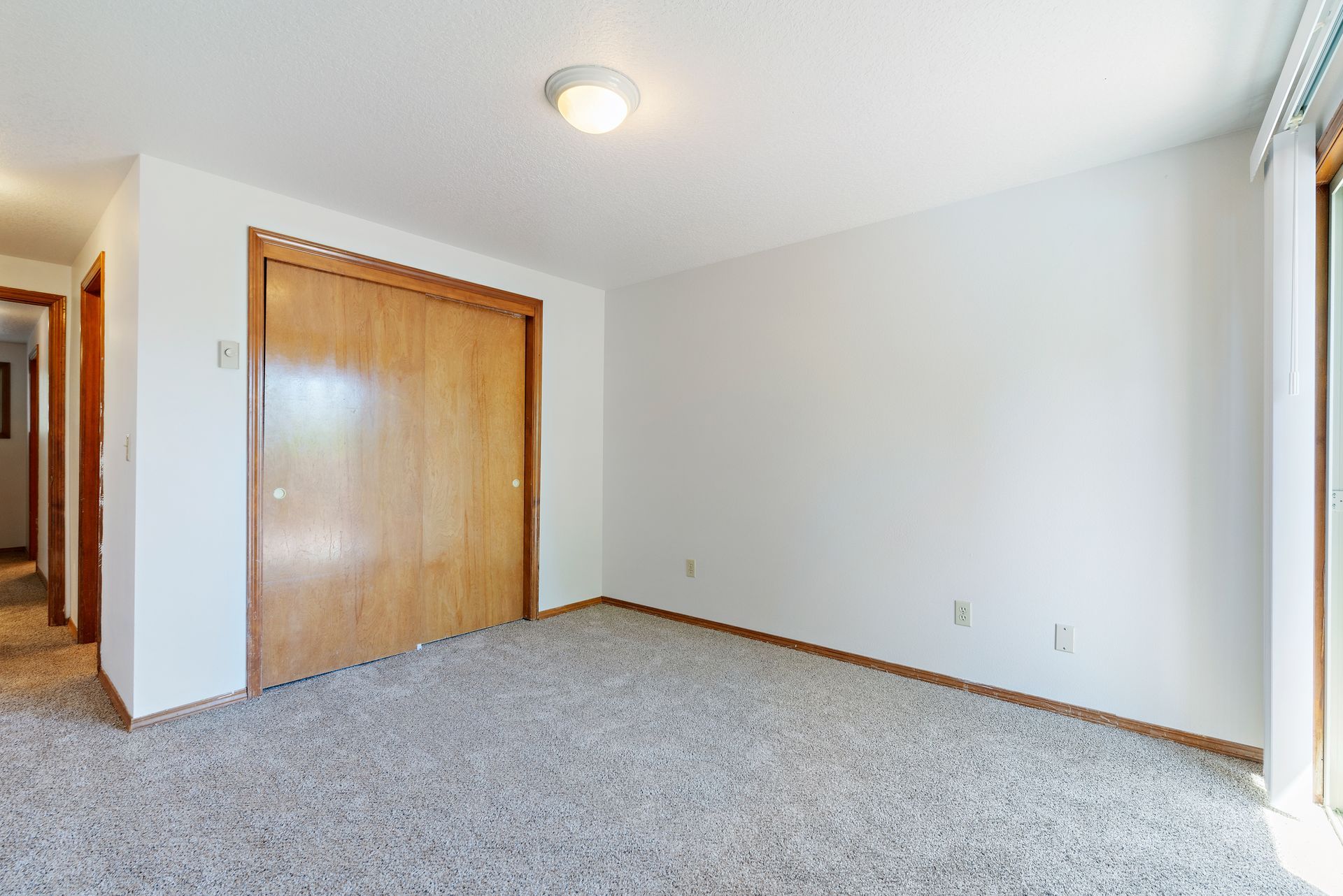 Empty carpeted bedroom with white walls, wood closet doors, and a window on the right.