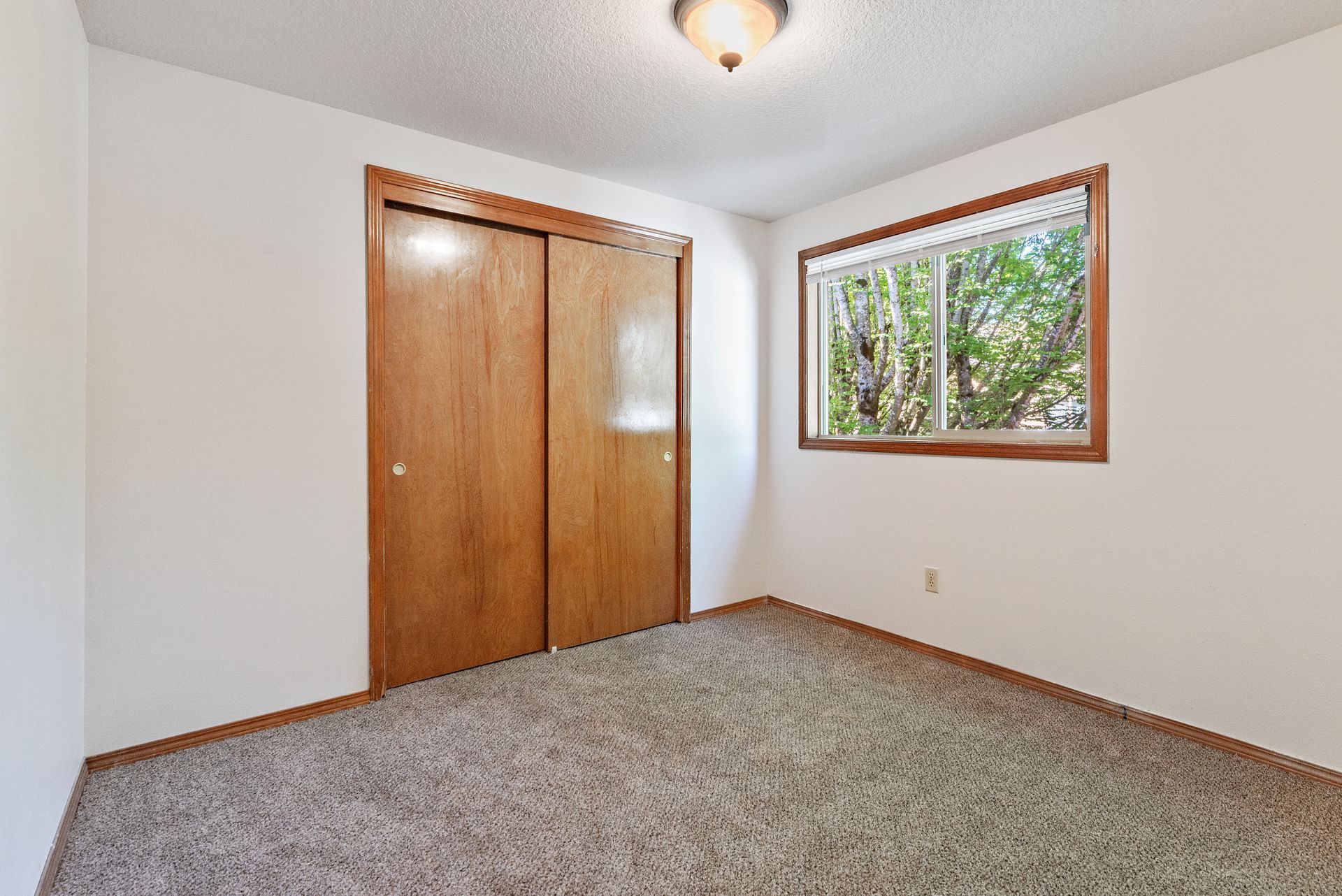 Empty bedroom with carpet, white walls, wooden closet doors, and a window looking out to trees.