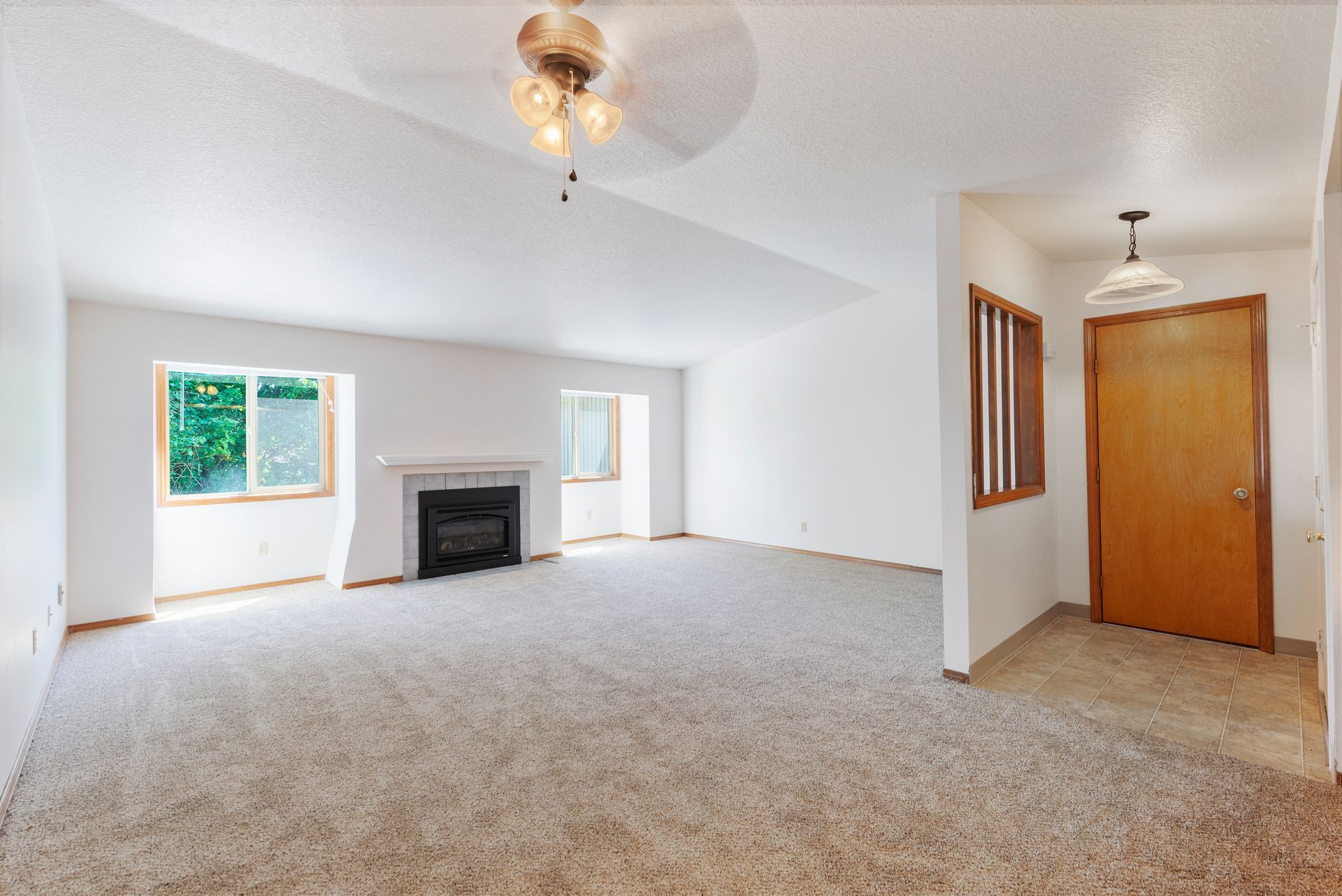 Empty carpeted living room with fireplace, ceiling fan, and doorway to hall with wooden closet doors