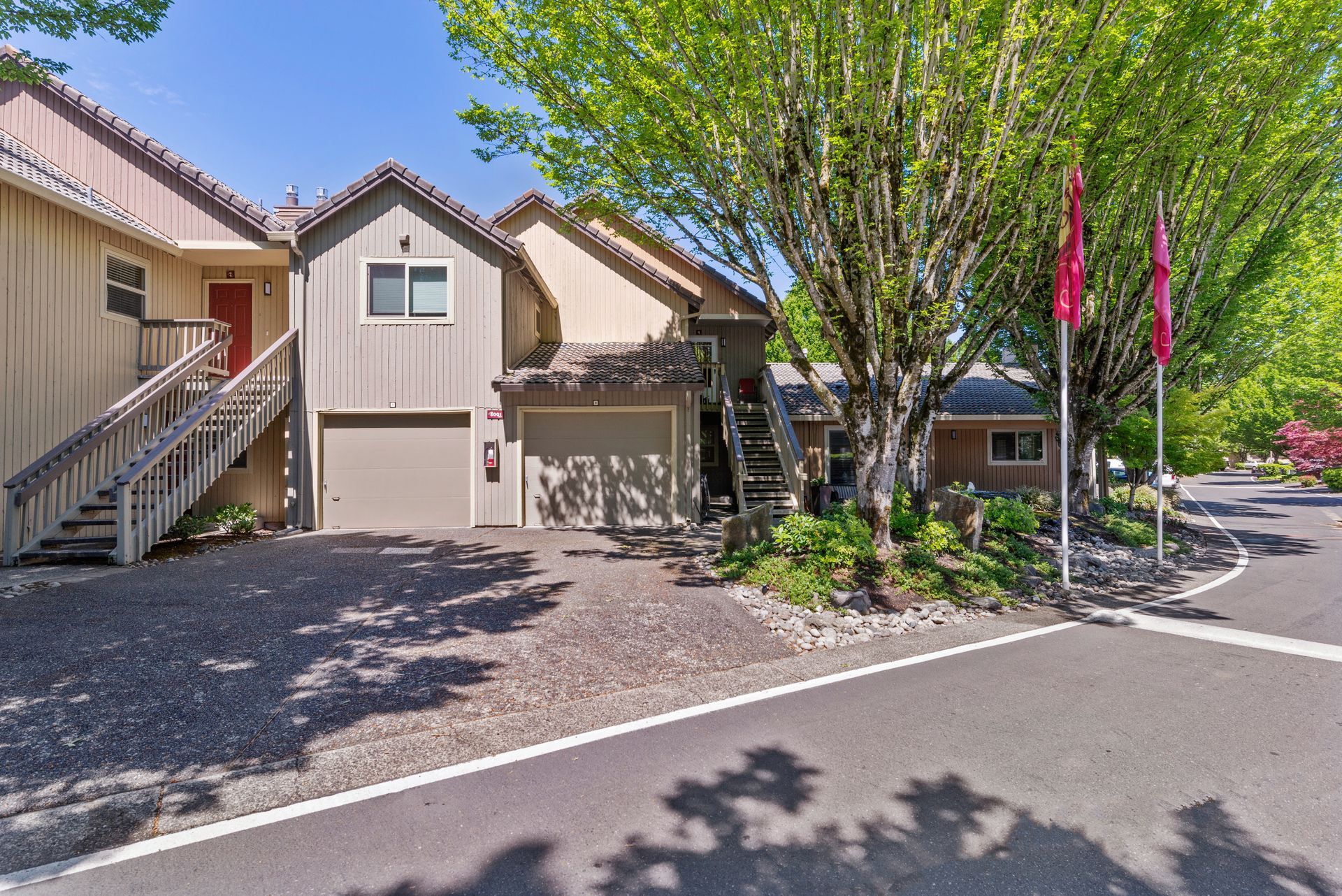 Row of tan townhomes with exterior stairs, trees, and pink flags along a sunny street