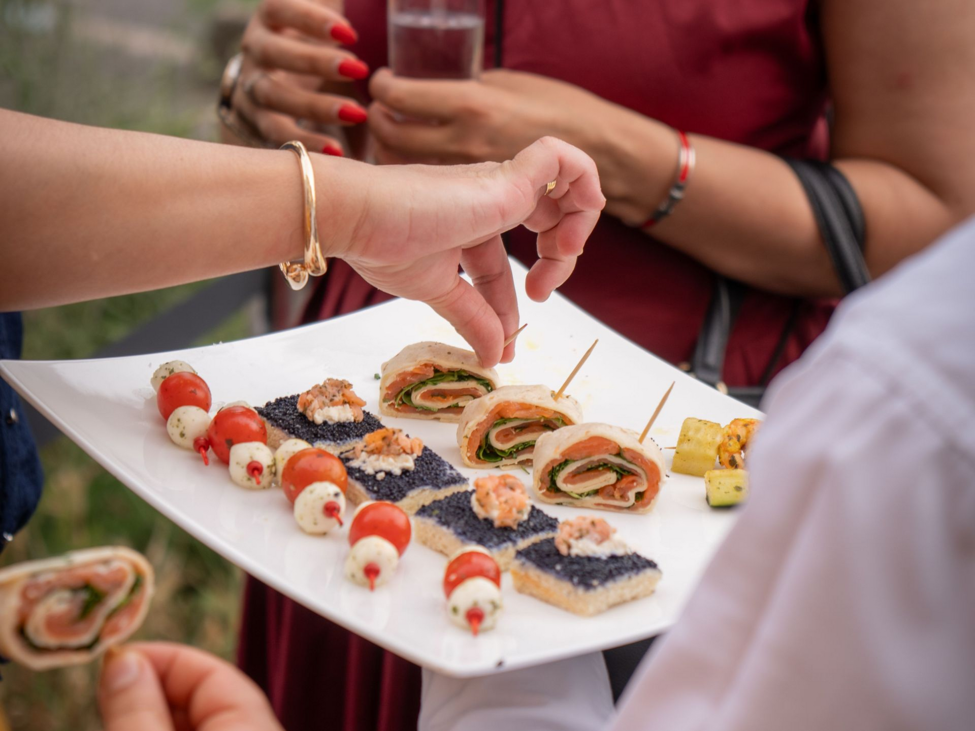 Hands selecting sushi appetizers from a white platter at a social gathering