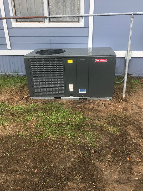 A black air conditioner is sitting in the dirt in front of a blue house.