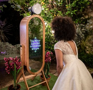 A woman in a wedding dress is standing in front of a mirror that says `` the mirror is excited to celebrate with you ''.