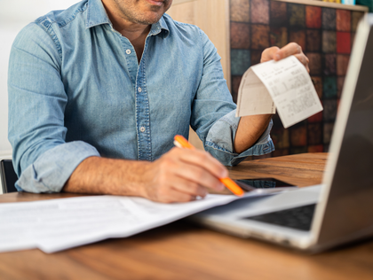 Person writing at a desk with a laptop and holding a receipt, surrounded by papers.