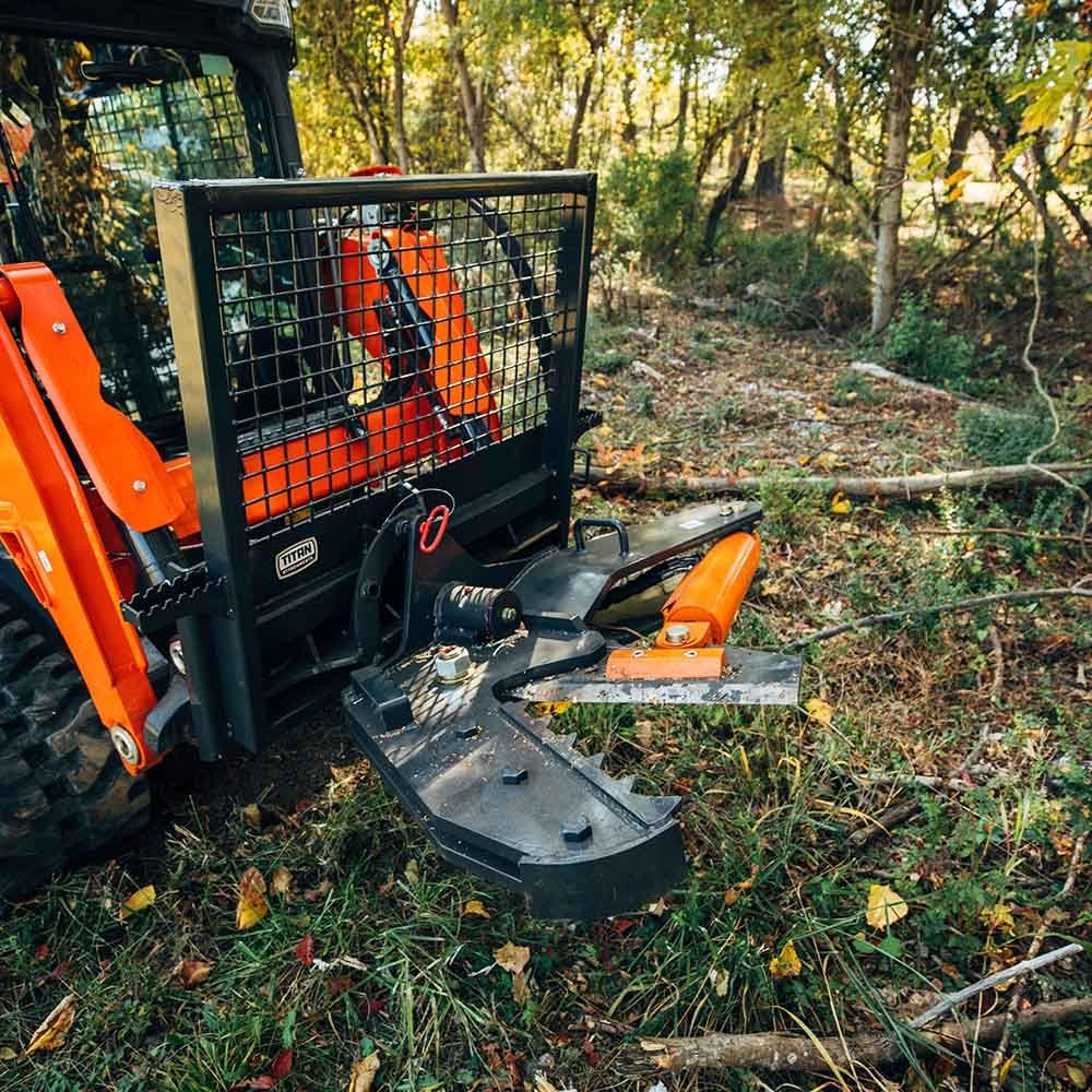 Close-up of a skid steer forestry grapple attachment in a forest setting, featuring orange and black metal components.