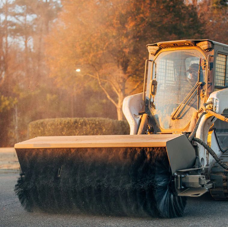 A skid-steer loader equipped with a large, rotating brush attachment for cleaning, set against a golden, sunset background.