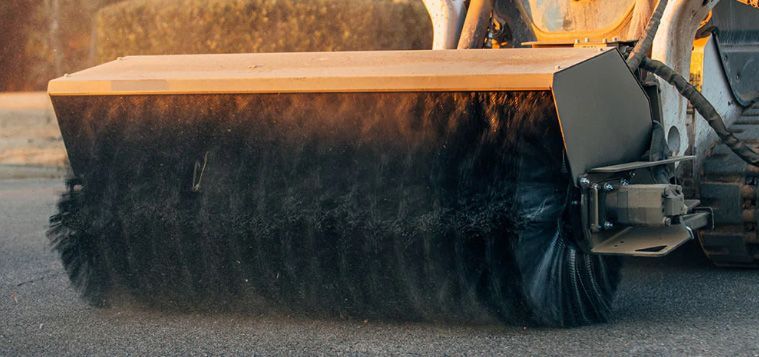 A black, cylindrical broom attachment mounted to the front of a skid-steer loader for sweeping paved surfaces.