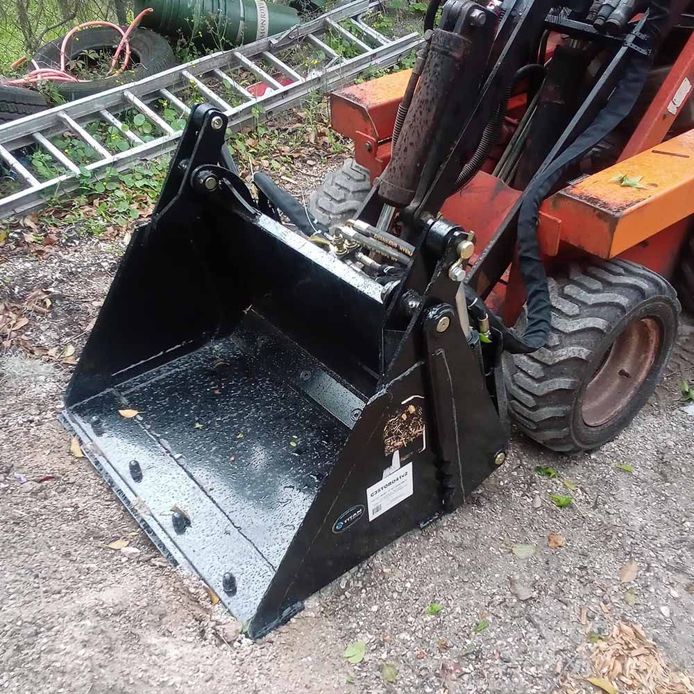 A shiny black hydraulic grapple bucket attached to an orange skid steer loader, sitting on a gravel surface outdoors.