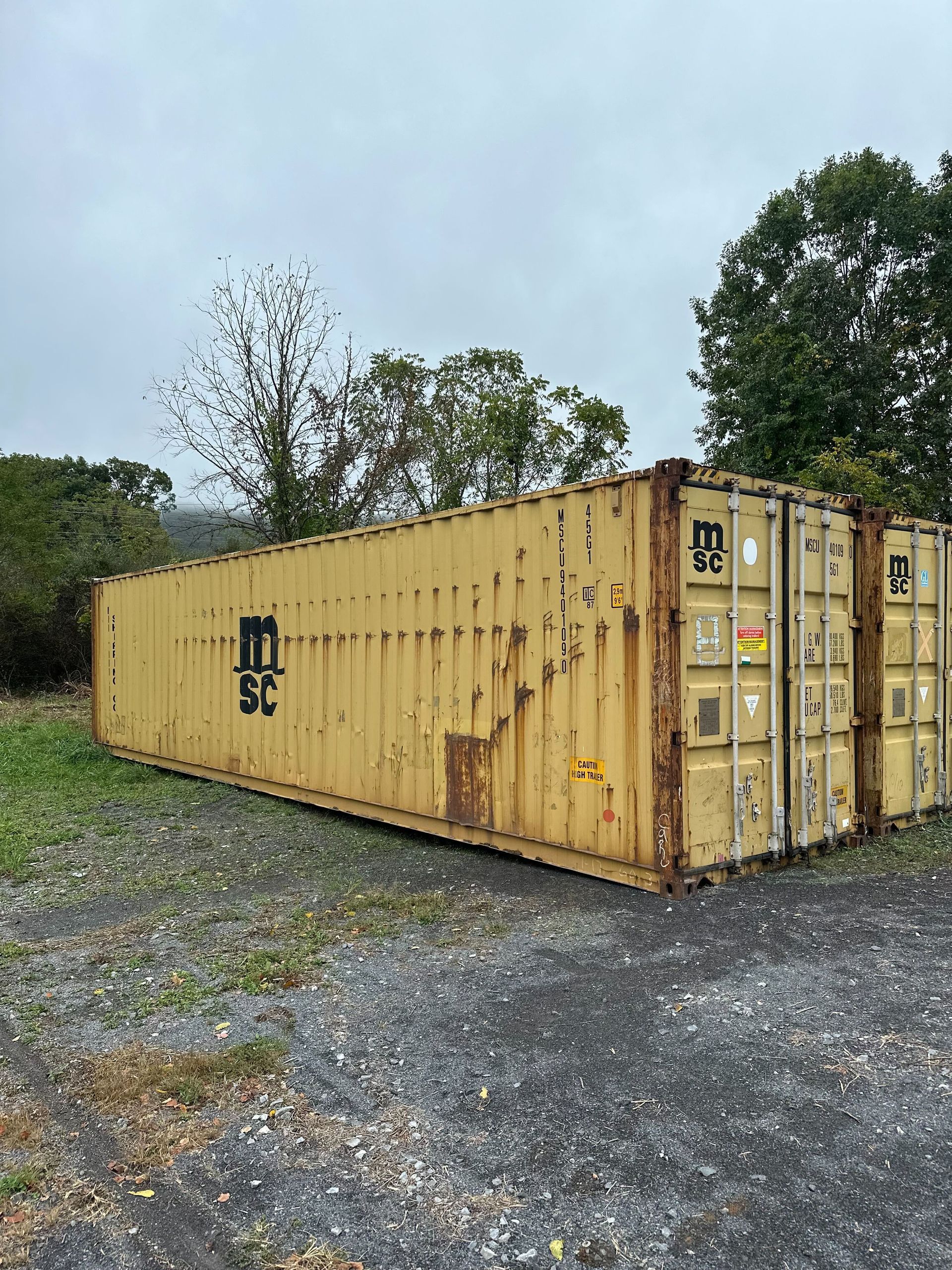 A tan, weathered MSC shipping container sits on a gravel lot under a cloudy sky, surrounded by trees.