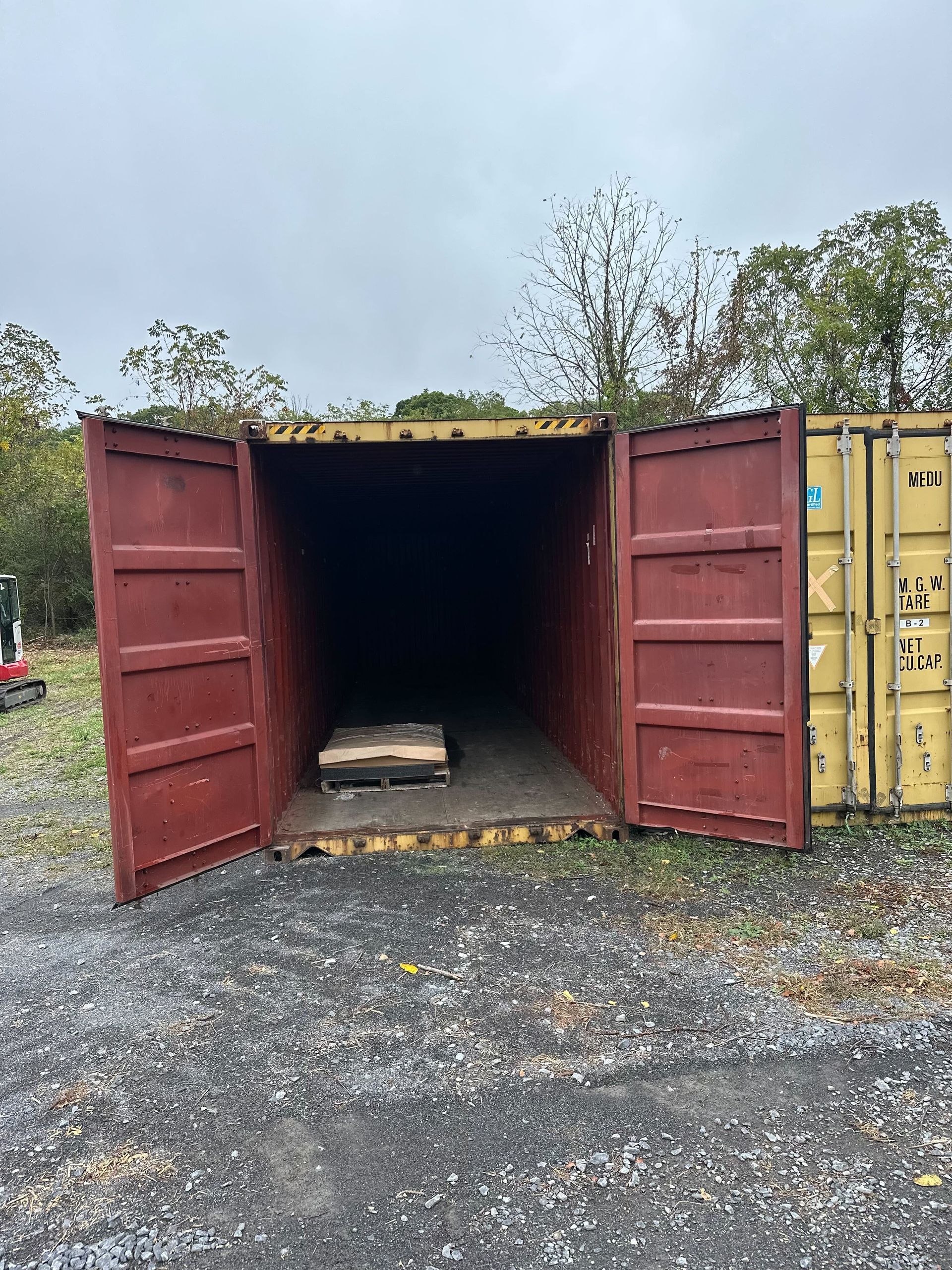 Open maroon shipping container on a gravel lot next to a yellow container, with a single pallet sitting inside.