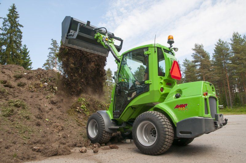 A bright green compact loader dumps a bucket of dirt onto a pile outdoors on a sunny day.