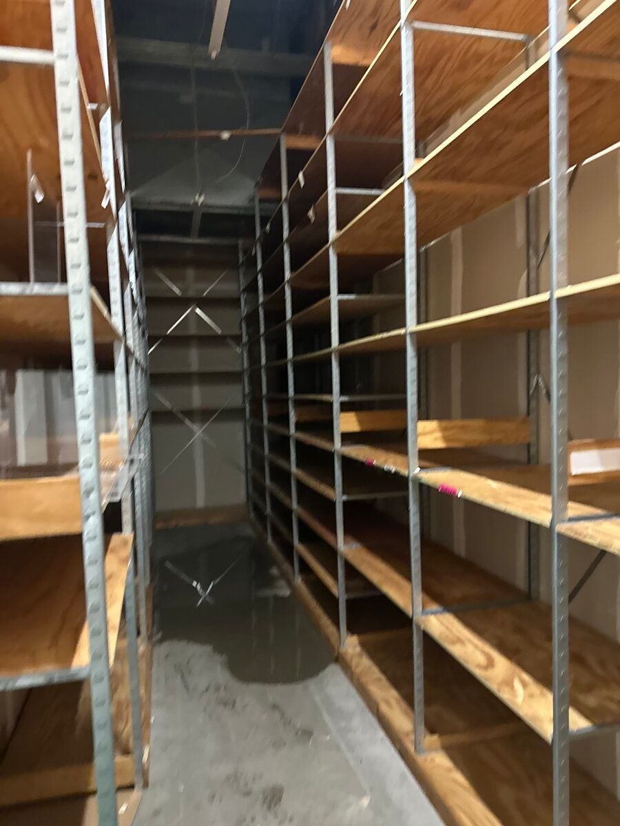 A narrow aisle between two rows of tall, multi-level metal shelving units with wooden shelves in a storage room.