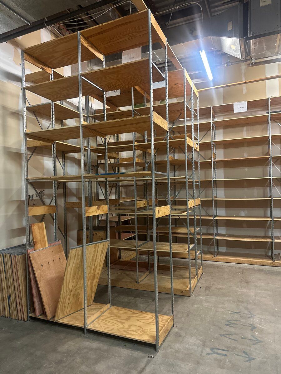 A tall metal shelving unit with wooden boards stands in a warehouse space next to a wall-mounted set of empty shelves.