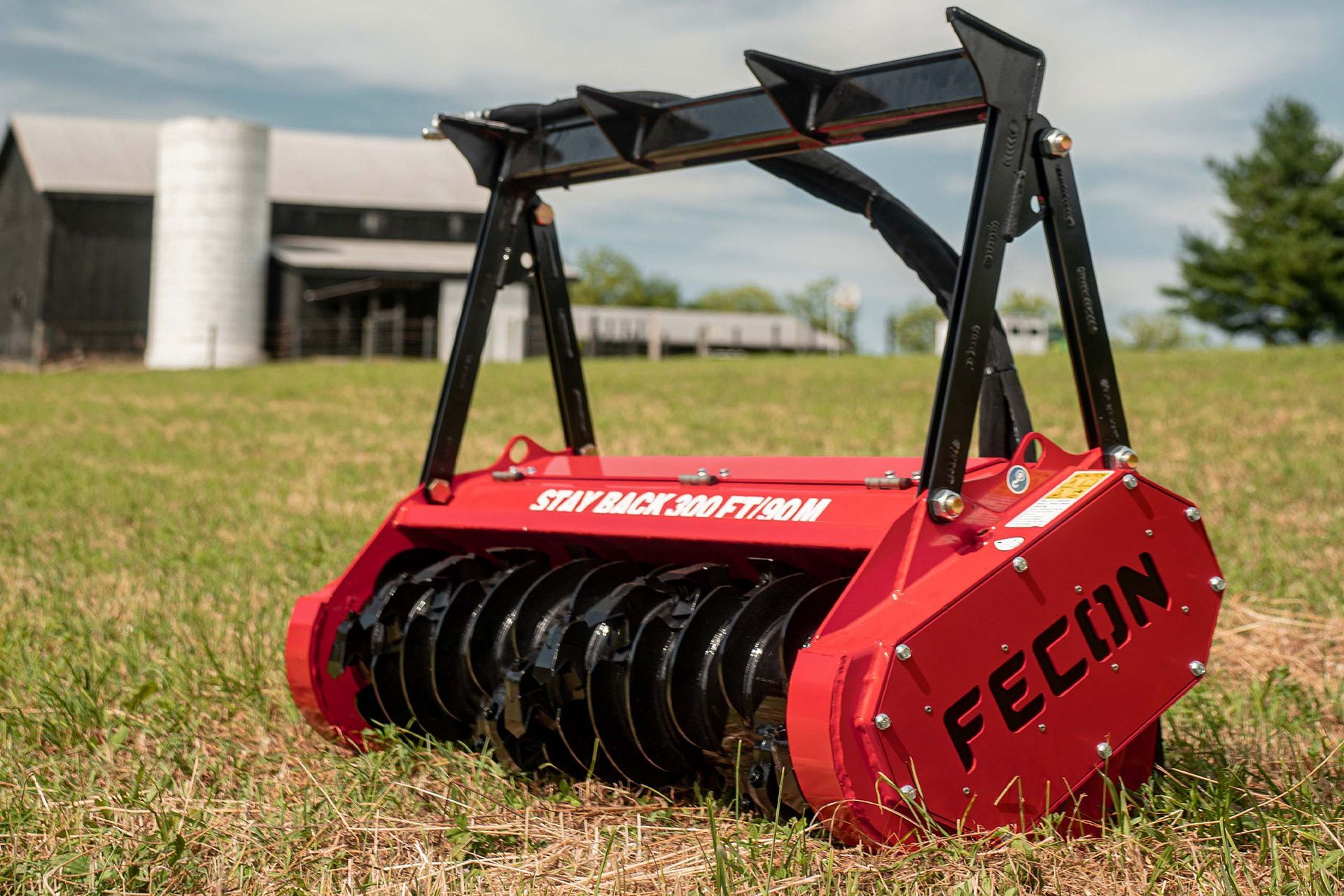 A bright red Fecon forestry mulcher attachment sits in a grassy field with a barn and silo in the background.