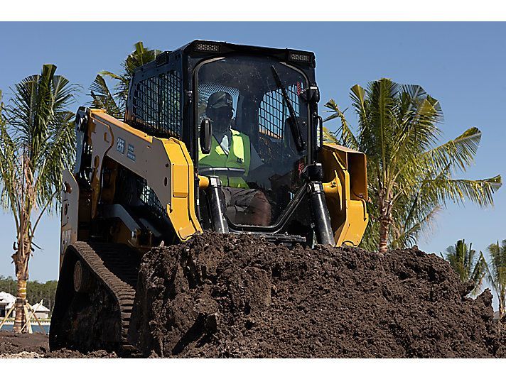 A yellow tracked skid steer loader moves a pile of dirt outdoors with palm trees in the background.