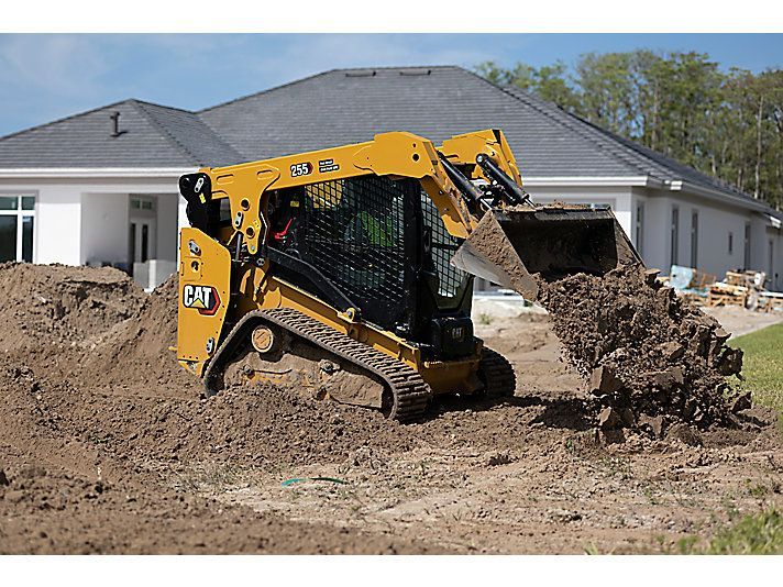 A yellow Caterpillar compact track loader moving dirt in front of a house under construction.