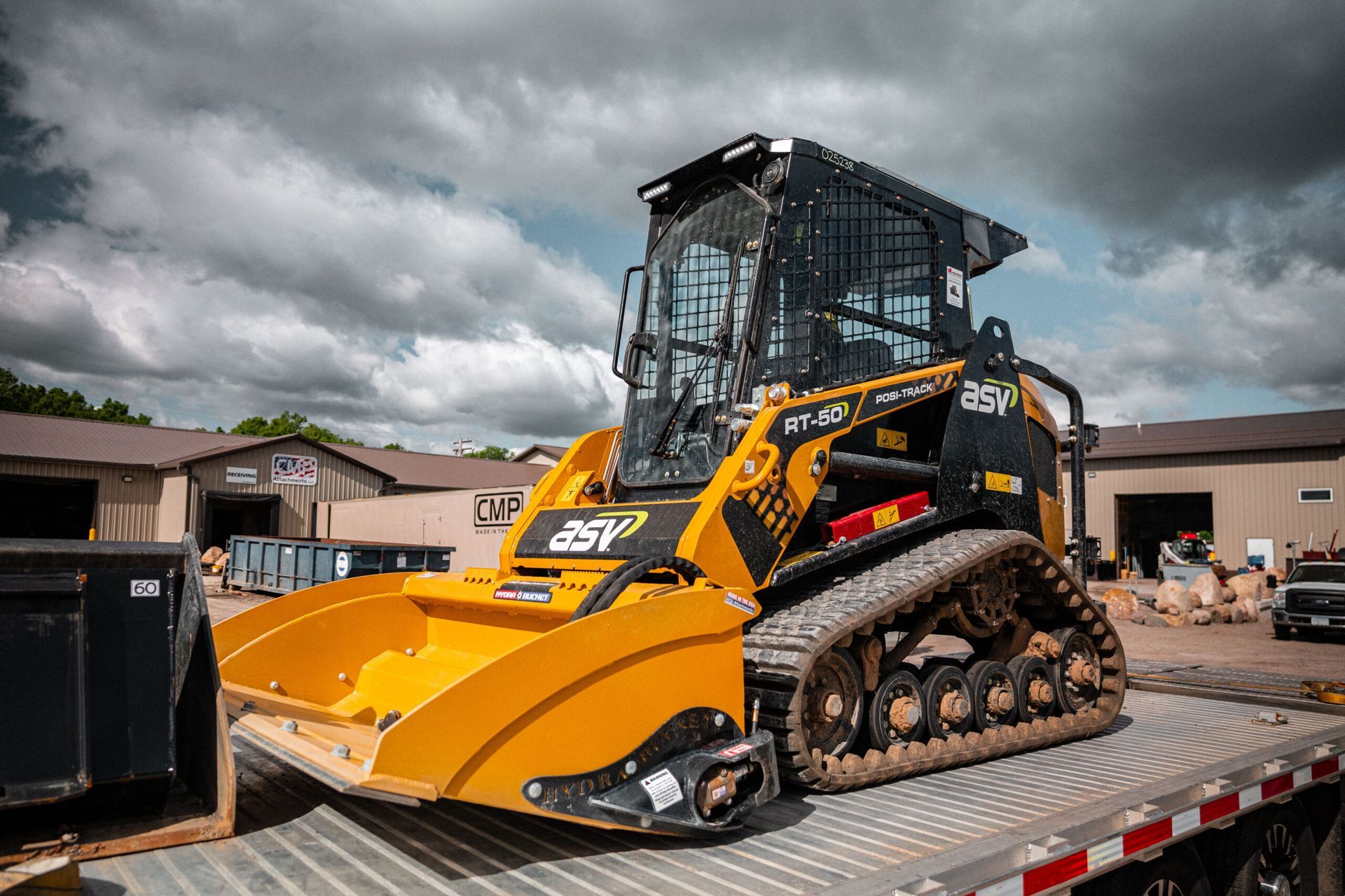 A yellow and black ASV compact track loader sits on a flatbed trailer outdoors under a cloudy sky.