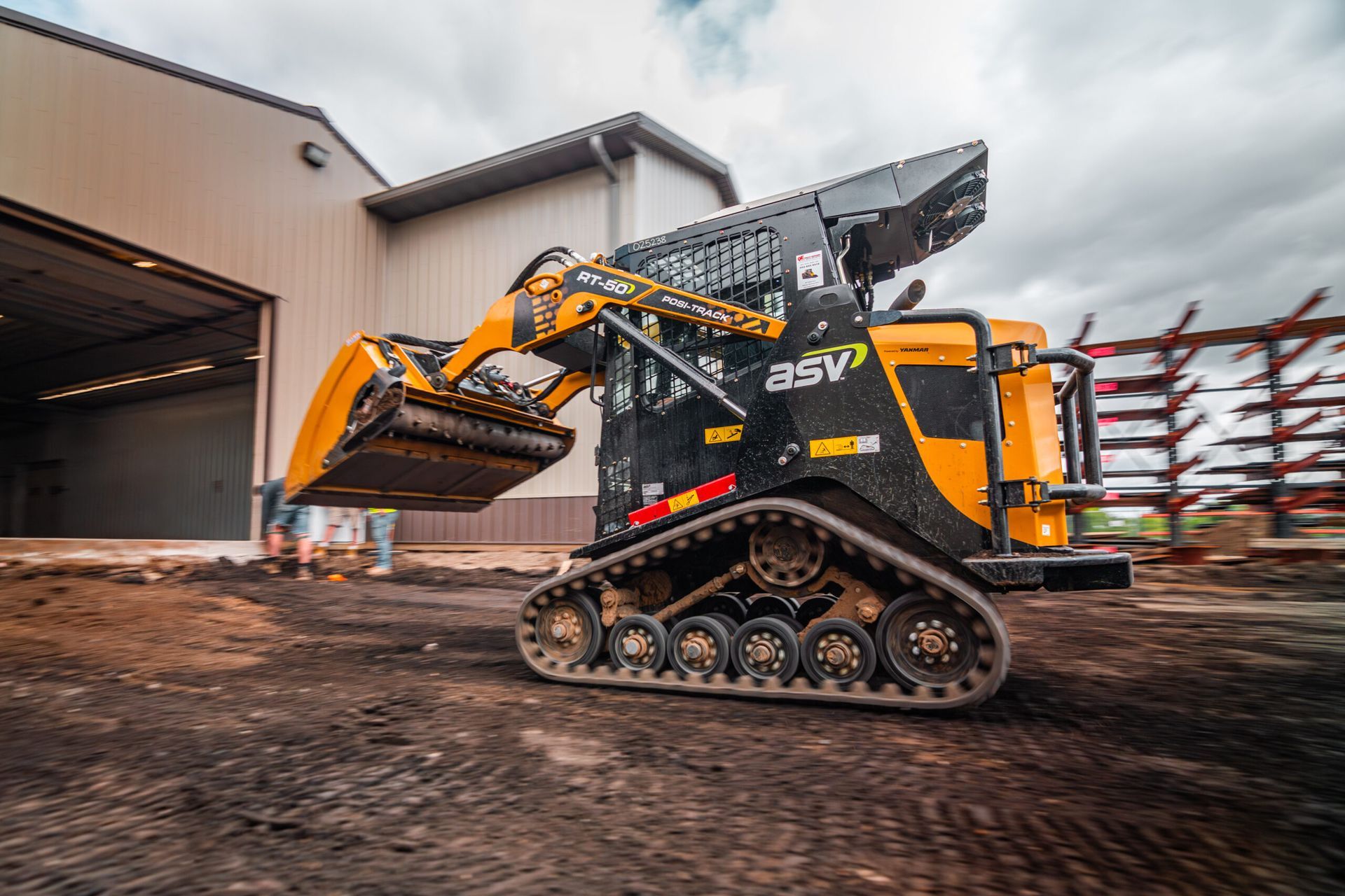 A yellow and black ASV compact track loader with a bucket attachment parked outside a large building on a dirt lot.