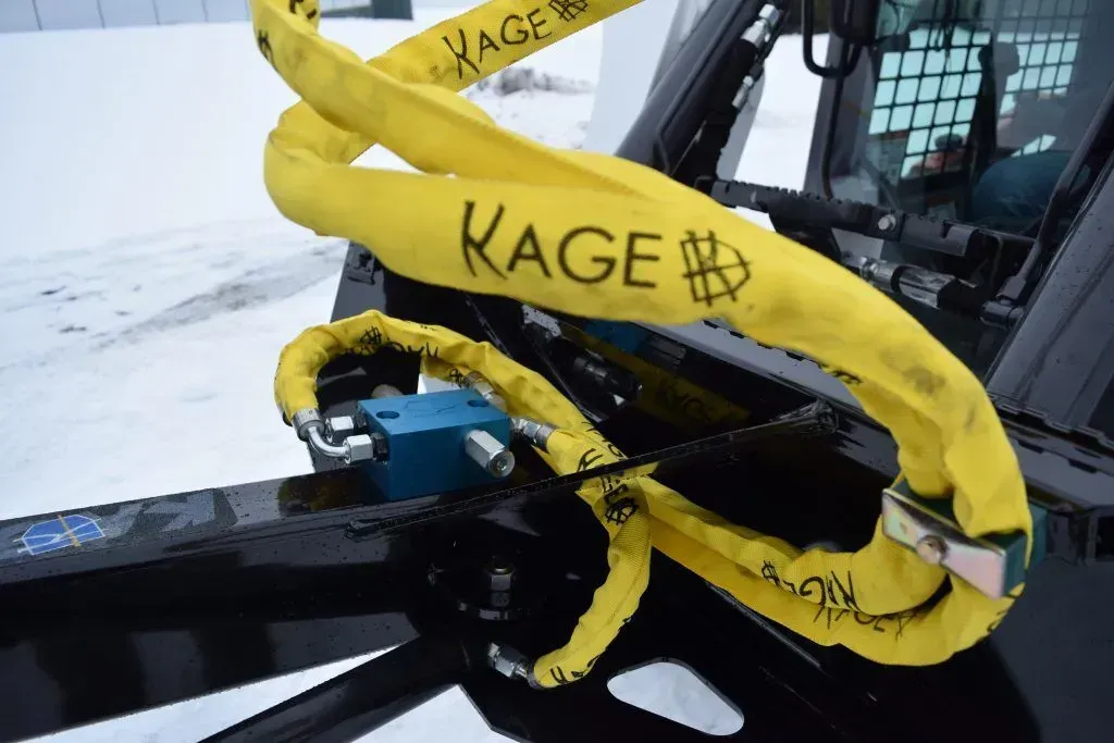 A yellow protective sleeve with the KAGE logo on a hydraulic line attached to heavy machinery in a snowy setting.