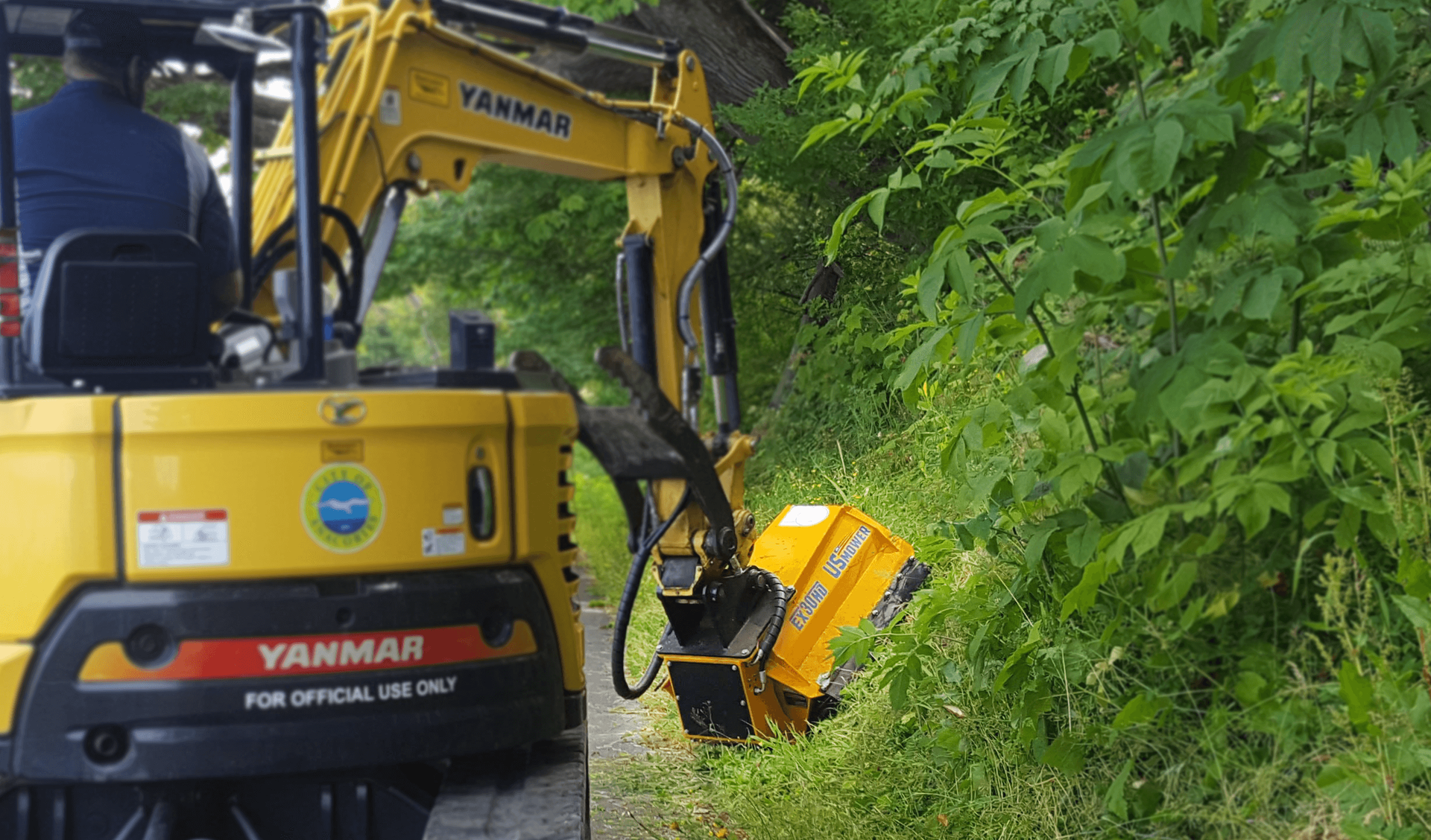 A yellow Yanmar mini-excavator with a brush cutter attachment clears vegetation along a wooded roadside.