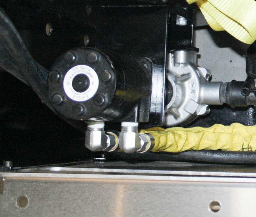 A black motor mounted to a metal bracket with two silver, right-angled pipe fittings and yellow hydraulic hoses.