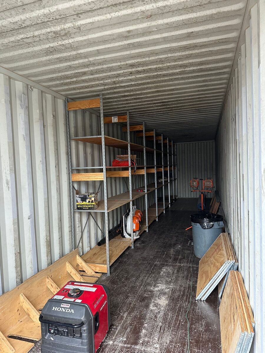 A shipping container interior with metal shelving on one side, a red portable generator, and wood planks on the floor.