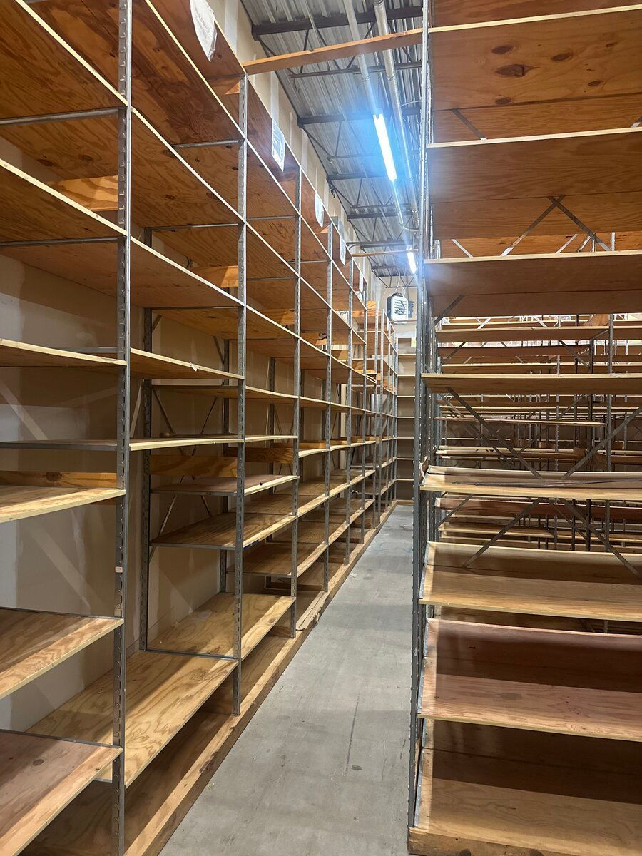 Empty industrial shelving units made of metal frames and plywood boards lining a narrow aisle in a storage area.