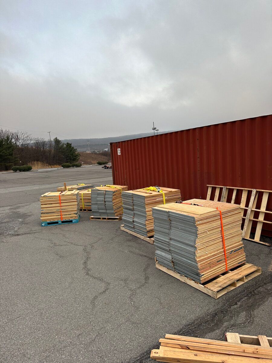 Stacked pallets of construction materials with orange straps sit on an asphalt lot beside a large red container.