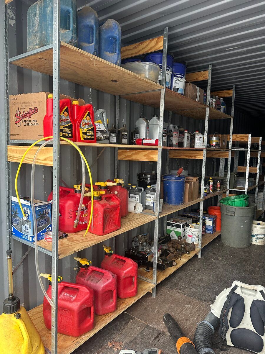 Metal storage shelving in a shipping container filled with red gasoline cans, bottles of oil, and miscellaneous supplies.
