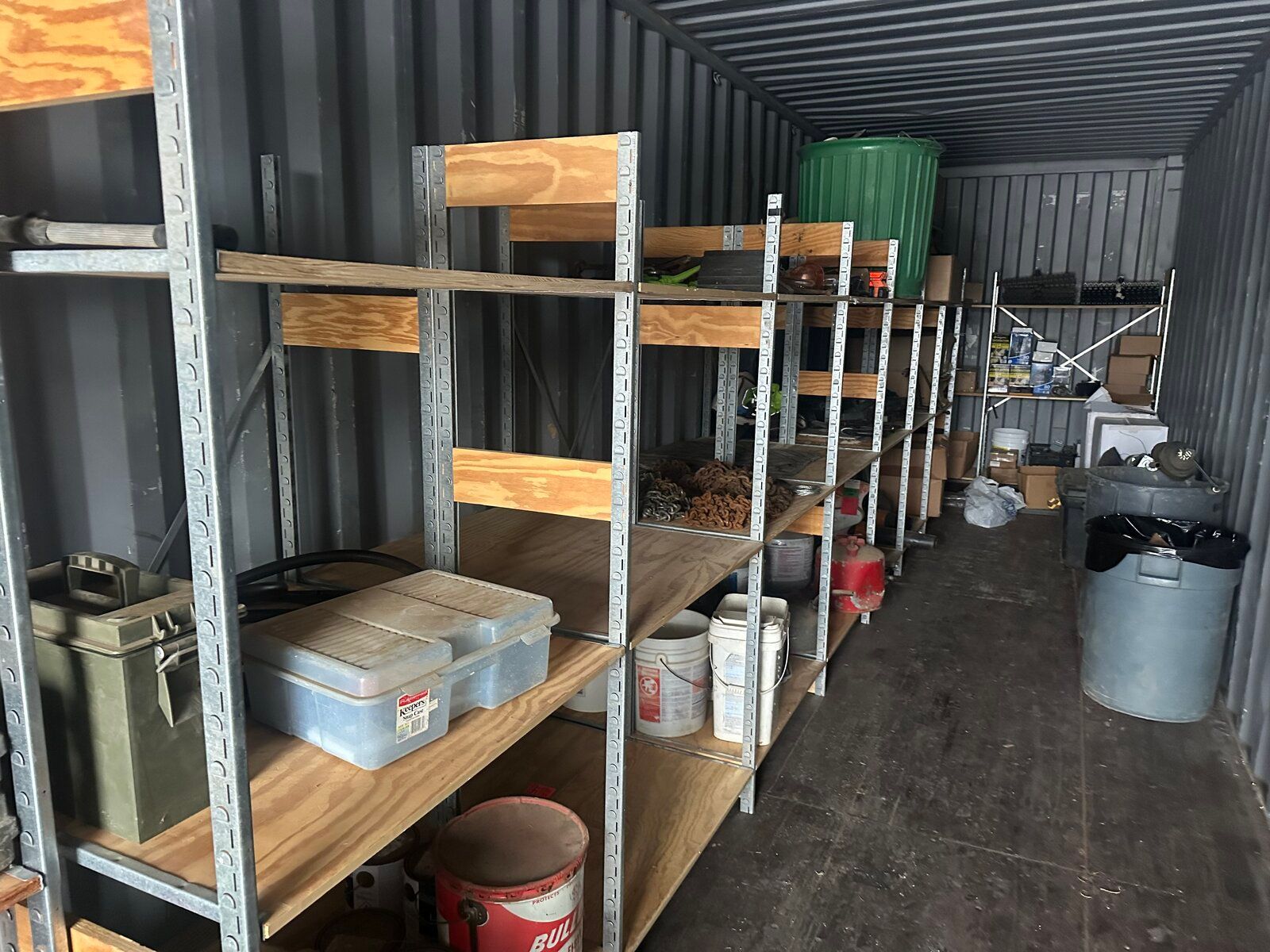 Storage shelves inside a long, narrow shipping container with various supplies, boxes, and bins.