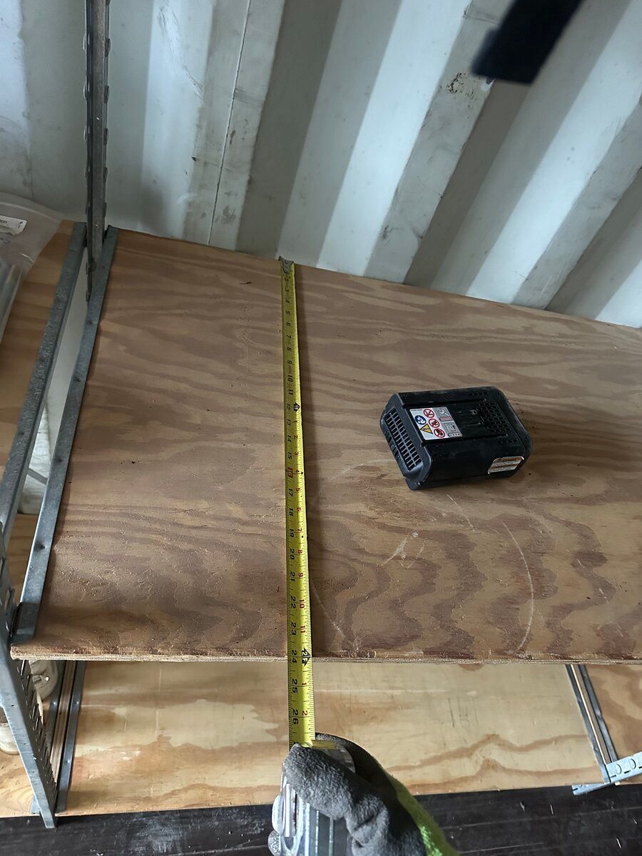 A gloved hand uses a yellow tape measure to determine the depth of a wooden shelf inside a shipping container.