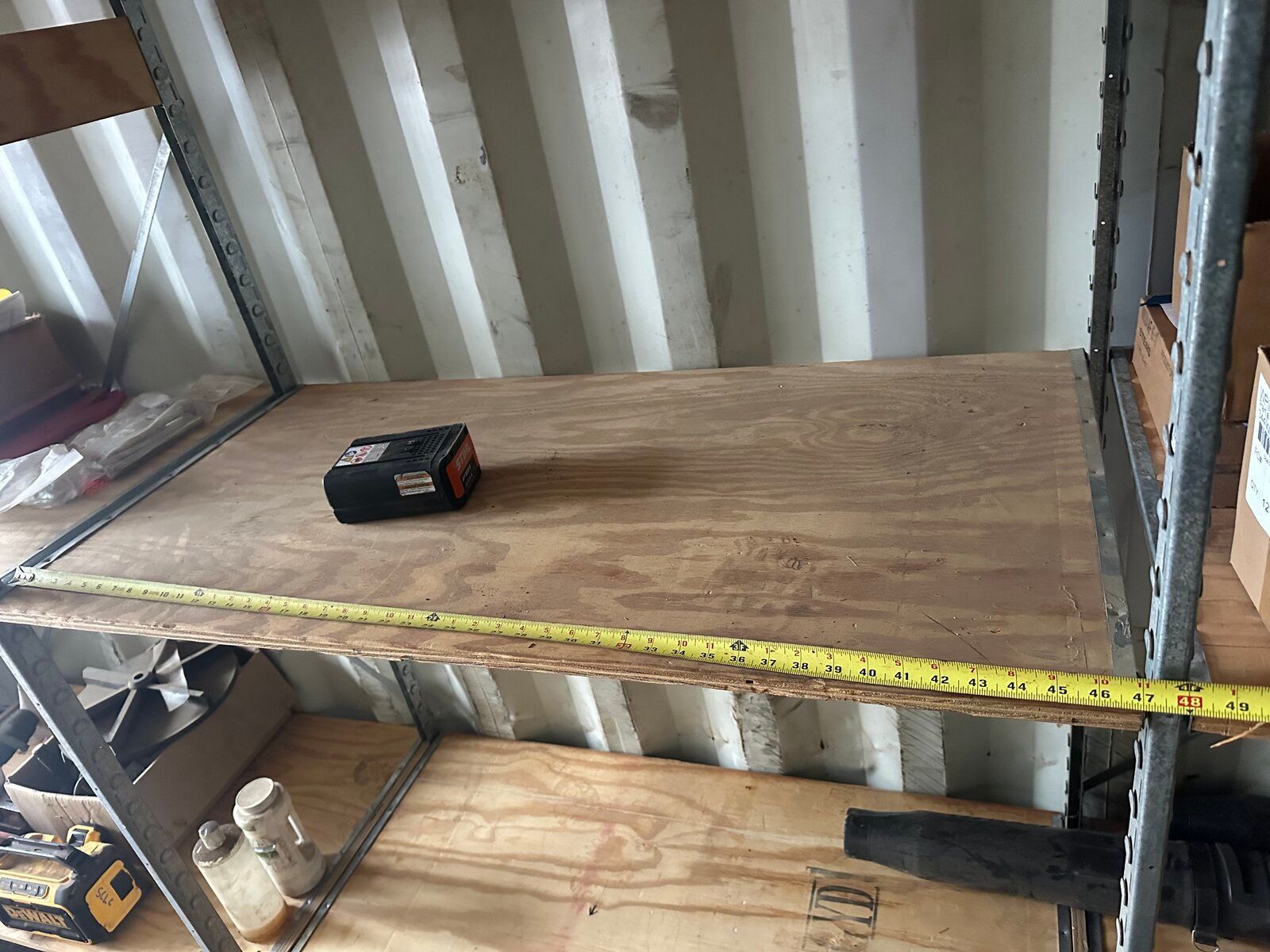 A yellow measuring tape extended across a wooden shelf inside a metal shipping container, with a battery pack on top.