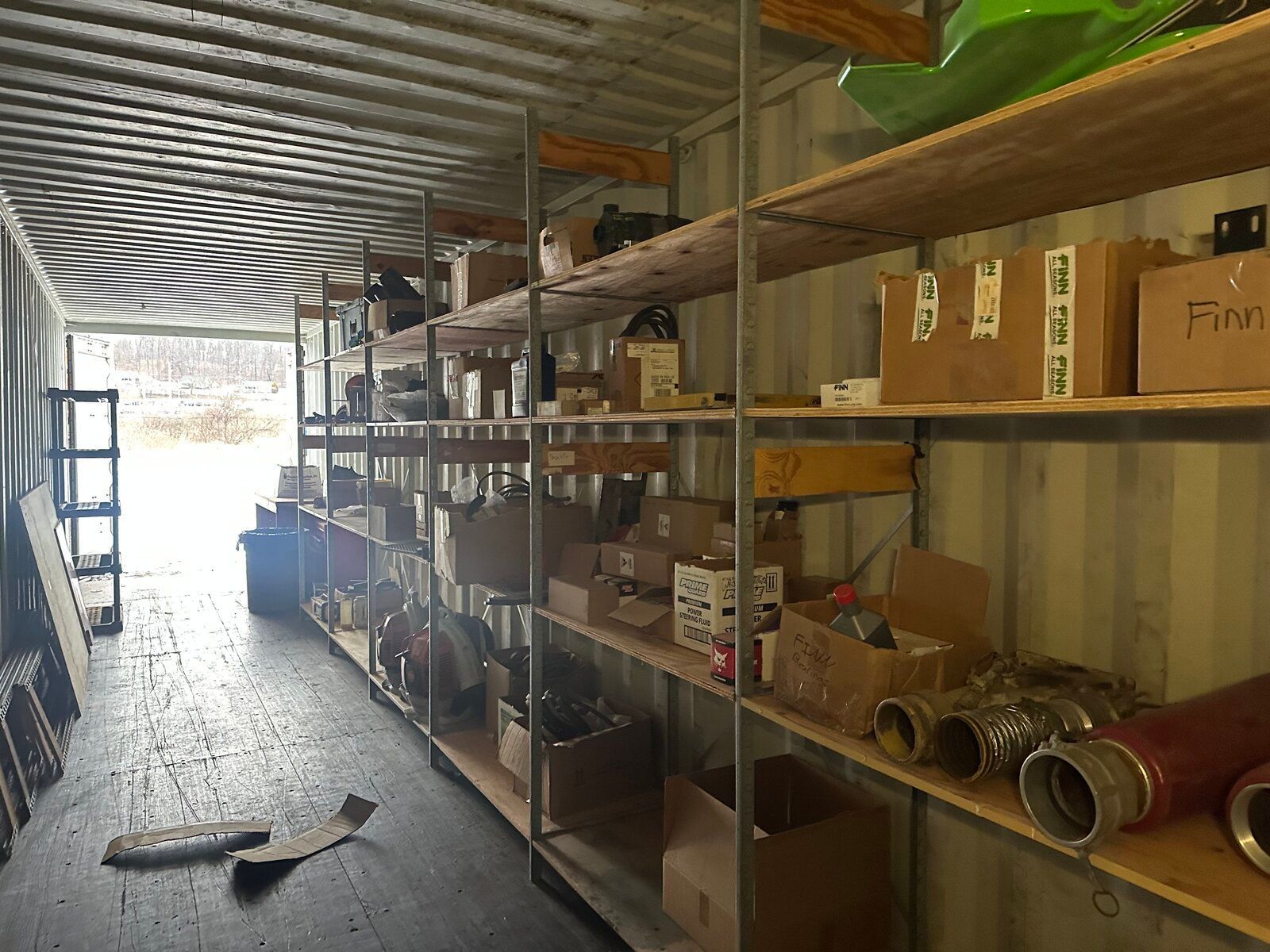 Inside a storage shipping container, long rows of metal shelving hold various cardboard boxes and industrial equipment.