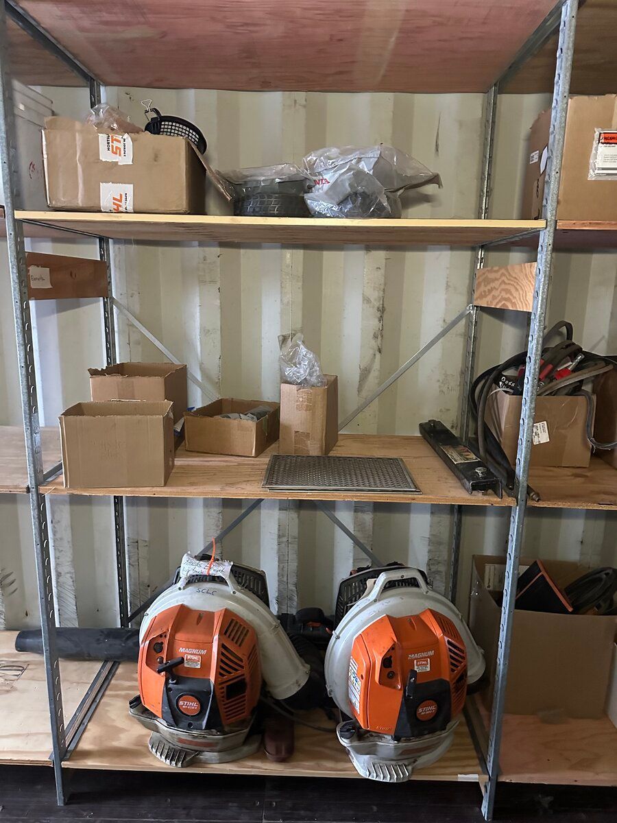 Metal shelving unit holding cardboard boxes and various equipment, with two orange leaf blowers on the bottom shelf.