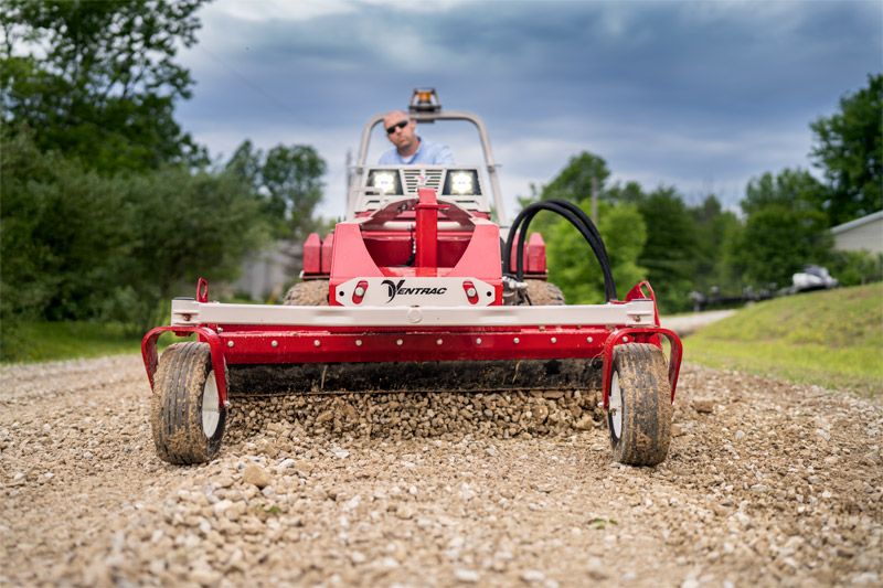 A person drives a red Yanco driveway grader on a gravel road under a cloudy sky.