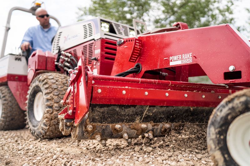 A person drives a red tractor equipped with a Power Rake attachment over a gravel surface.