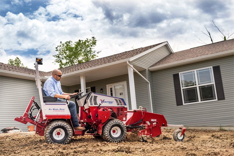 A person operates a red and white Vermeer utility tractor to grade soil in front of a residential house.