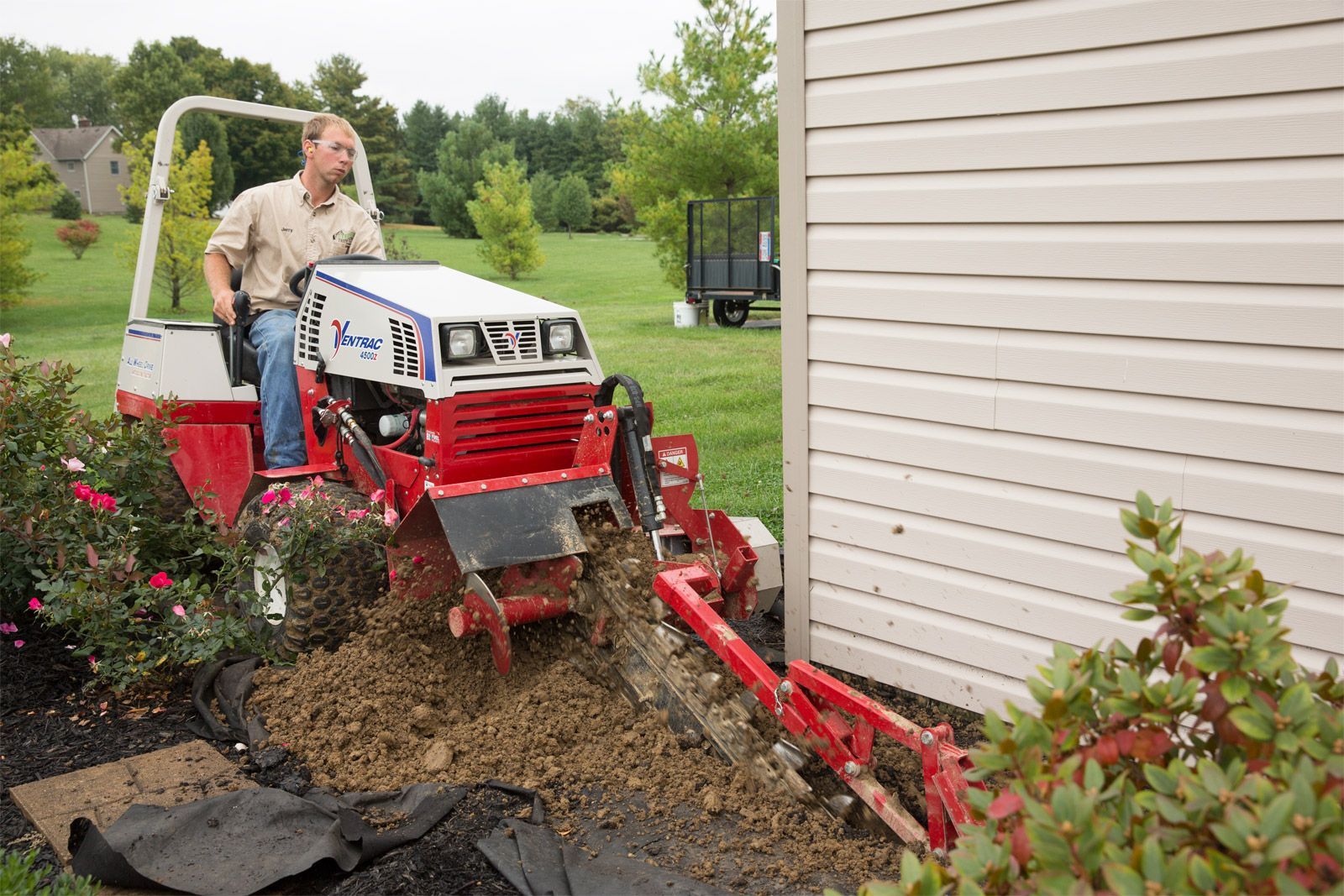 A person operates a red and white trencher machine to dig a trench along the side of a house in a grassy yard.