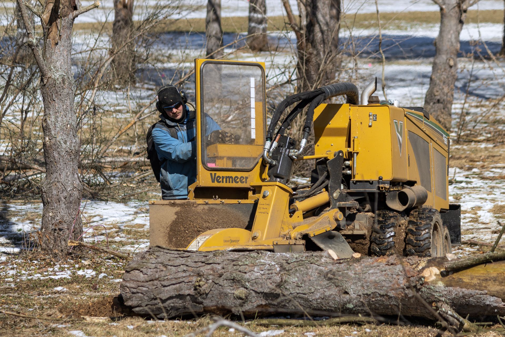 A person wearing hearing protection operates a yellow Vermeer stump grinder on a snowy, wooded lot.