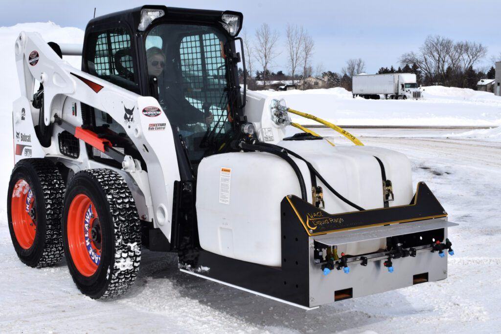 A white Bobcat skid-steer loader equipped with a large liquid storage tank and sprayer attachment on a snowy lot.