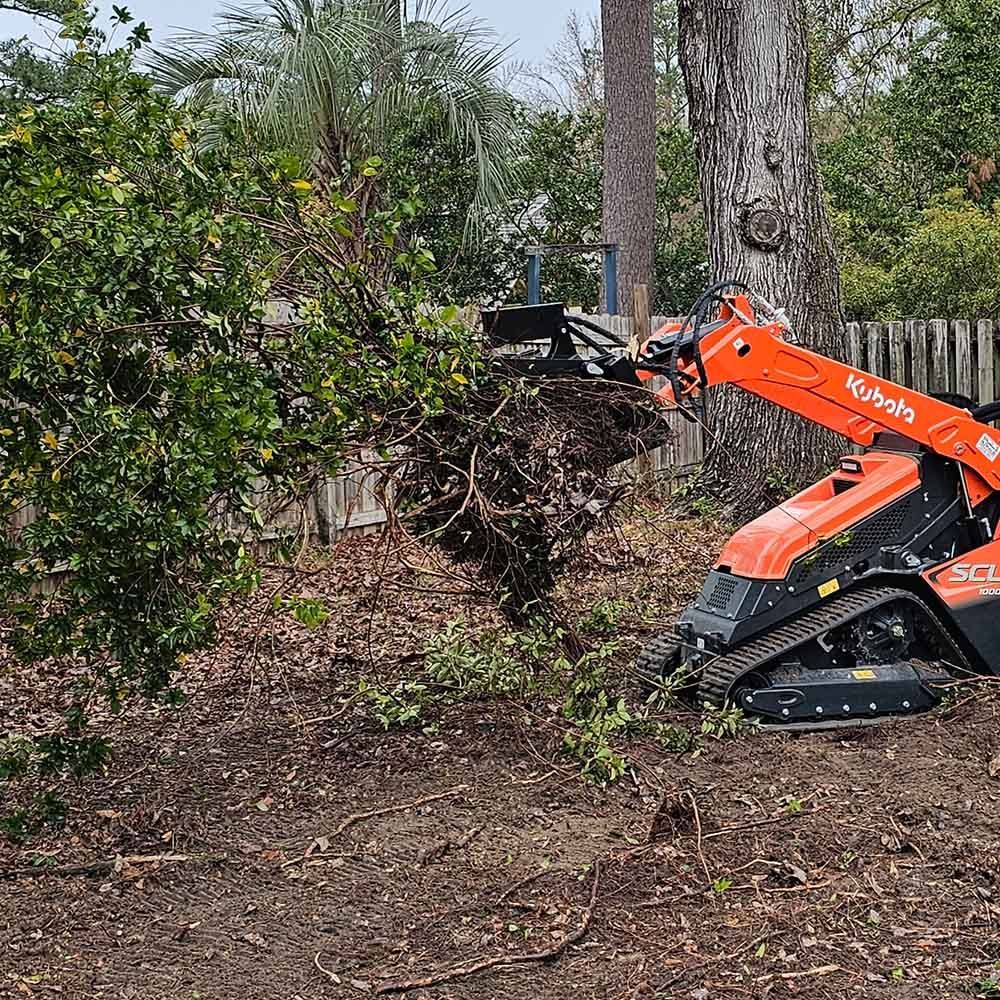 An orange compact track loader with a grapple attachment moves a pile of brush in a backyard.